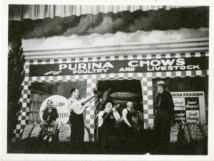 A black-and-white photograph of the Grand Ole Opry stage, adorned with sponsor Purina's name, a checkerboard motif, and several performers gathered around Uncle Dave Macon.