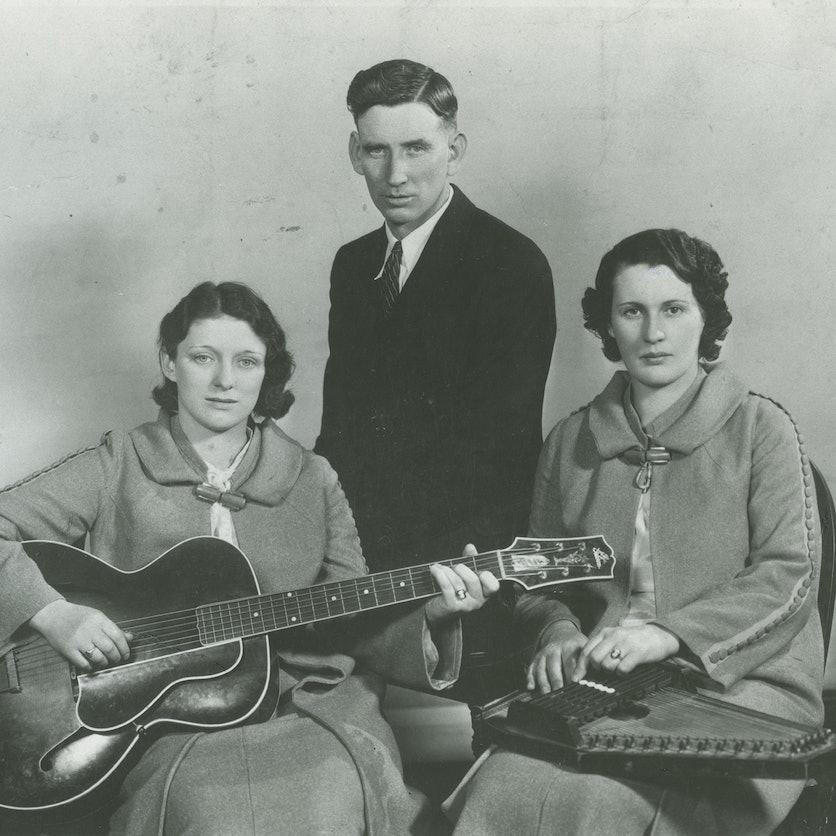 Three members of the Carter Family pose for a promotion photo taken in black and white. Maybelle Carter is seated holding a guitar and facing the camera. She is wearing a unique coat with a wooden clasp at the collar. Seated beside her is Sara Carter - she is holding an auto harp instrument in her lap and pressing the keys. She is wearing the same outfit as Maybelle, a unique coat with a wooden clasp at the collar. Leaning behind them is A.P. Carter, he is wearing a dark suit jacket and tie.