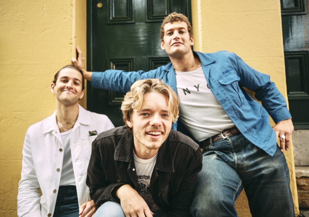 Three young men are posing and smiling for a promotion image of their band. The man on the far left is smiling and wearing a white button up shirt and jeans. The man in the middle is wearing a brown shirt and jeans and leaning down towards the camera, his hair is in his face. The man on the far right is leaning against a yellow wall behind the other two men, and wearing a blue button up shirt with a white tee shirt with the text “NYC” on the shirt. 